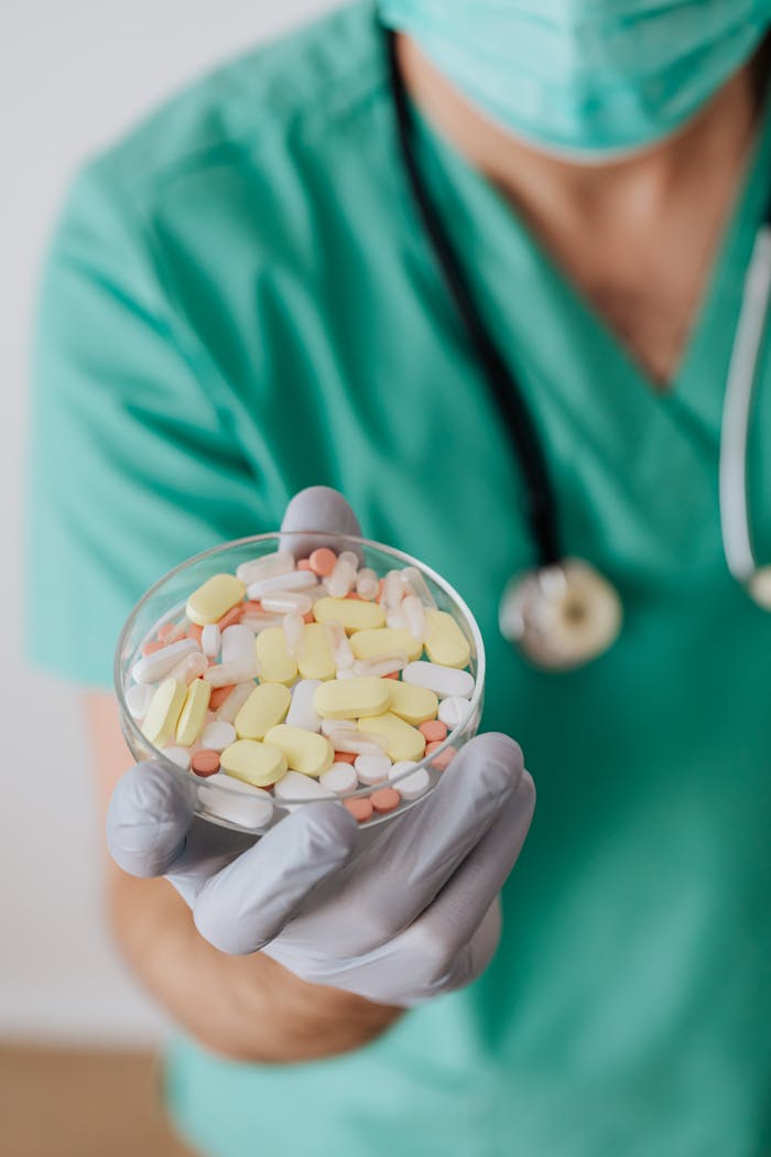 Unrecognizable male doctor wearing medical uniform and mask and gloves showing different medicine during work standing with stethoscope near white wall blurred background